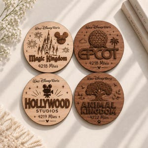 Op de afbeelding: Vier ronde houten onderzetters met gegraveerde ontwerpen van Walt Disney World-parken. De onderzetters tonen Magic Kingdom, Epcot, Hollywood Studios en Animal Kingdom met parknamen, afstanden in mijlen en iconen zoals een kasteel, bol, camera en boom.
