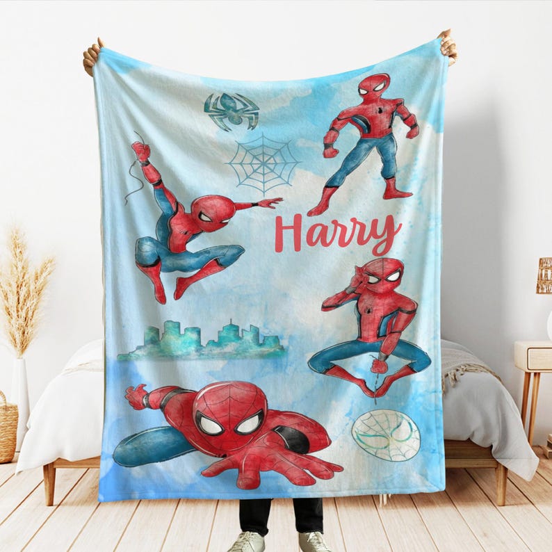 K&ouml;nnte beinhalten: Eine hellblaue Decke mit Illustrationen von Spider-Man in verschiedenen Posen. Das Design umfasst eine Stadtlandschaft, ein Spinnennetz und den Namen "Harry" in Rot. Die Decke hat einen Aquarellstil mit einem Spinnenlogo.