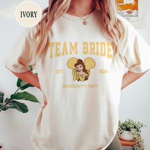 May include: Cream-coloured t-shirt featuring the text "TEAM BRIDE" in yellow block letters. Below the text is a graphic of a princess in a yellow dress with mouse ears. The text "EST. 2026 BACHELORETTE PARTY" is printed below the graphic.