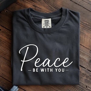 May include: Dark gray t-shirt with the word "Peace" in a white script font, and the phrase "- BE WITH YOU -" below it. The shirt is folded neatly on a wooden surface.
