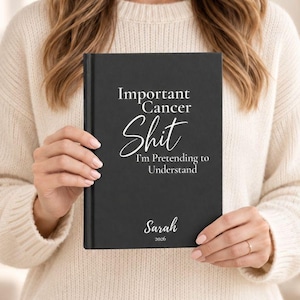 May include: A black hardcover journal featuring white text that reads Important Cancer Shit I'm Pretending to Understand, with the name Sarah and the year 2026 printed in white at the bottom center of the front cover.