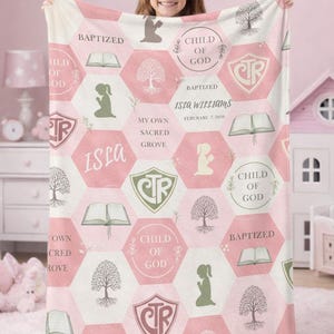 May include: A pink and white blanket with a hexagonal pattern. The blanket features text that says "Baptized", "Child of God", and "Isla Williams". It also has illustrations of a tree, a book, and a praying figure.
