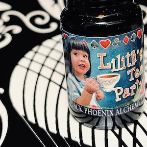 May include: A small, dark glass bottle with a black cap. The label features a child holding a teacup, with the text "Lilith's Tea Party" and "Black Phoenix Alchemy Lab". The background is black and white.