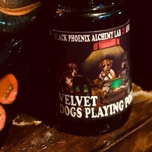 May include: A small, black bottle with a black cap. The label reads "BLACK PHOENIX ALCHEMY LAB" and features an illustration of dogs playing poker. The text "VELVET DOGS PLAYING POKER" is below the illustration. The bottle is on a wooden shelf.