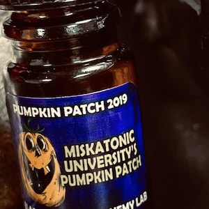 May include: A small, brown glass bottle with a black cap. The label reads "Pumpkin Patch 2019" and "Miskatonic University's Pumpkin Patch" with a cartoon pumpkin. The bottle is from Black Phoenix Alchemy Lab.