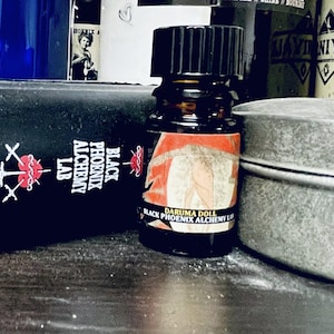 May include: A small, dark brown glass bottle with a black cap. The label features a red and white design with the text "Daruma Doll" and "Black Phoenix Alchemy Lab". The bottle is surrounded by other containers.