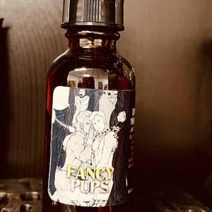 Puede incluir: Un frasco cuentagotas de vidrio ámbar oscuro con tapa negra. La etiqueta presenta una ilustración en blanco y negro de dos figuras y el texto FANCY PUPS en letras amarillas. El frasco es cilíndrico con cuello cónico y tapa de goma negra.