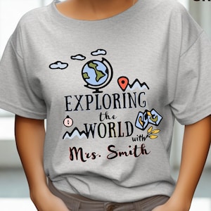 May include: A light gray t-shirt with the words "EXPLORING the WORLD with Mrs. Smith" in black script. The shirt features a globe, clouds, and map graphics in blue, red, and yellow. The words "SPORT GREY" are printed in the upper right corner.