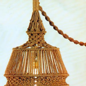 May include: A handcrafted macrame pendant light fixture. The light fixture is made of natural-colored rope, featuring a cylindrical shade with vertical strands and a decorative tassel. The fixture is suspended from a matching rope.