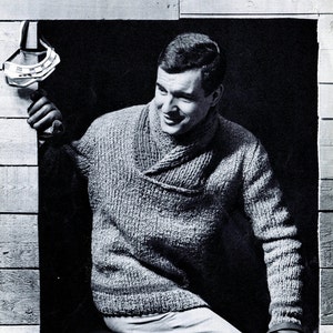May include: A man wearing a gray knitted sweater with a shawl collar, standing in a doorway. He is wearing gloves and looking to the side.