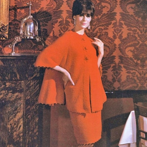 May include: A woman wearing a red dress with a matching cape. The dress has a scalloped hem and the cape has a scalloped edge. The woman is standing in front of a dark wood cabinet and a white tablecloth.