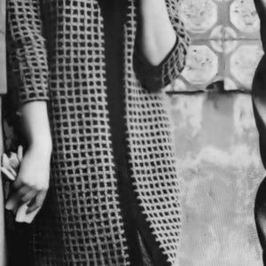 May include: A black and white photograph of a woman wearing a long coat with a grid pattern. The coat has three-quarter sleeves and a dark trim. She is smiling and holding a small object.
