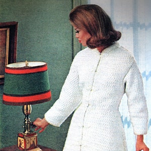 May include: A woman wearing a white crocheted robe with a button-up front. She is standing in a room with a wooden table and a lamp with a green and red striped shade.