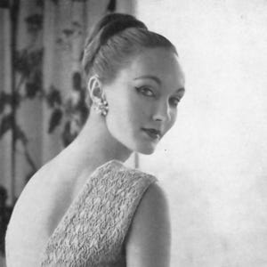 May include: A monochrome image of a woman in a textured dress with a single shoulder strap. The dress is belted at the waist. She wears a bracelet and earrings, with her hair styled up. A classic vintage look.