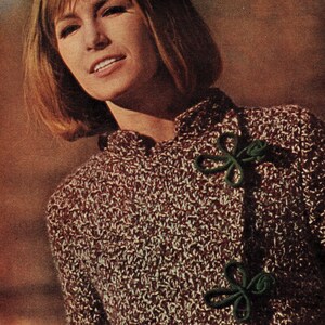 May include: A brown and white knit cardigan with green fabric flower embellishments. The cardigan has a high collar and a long sleeve.