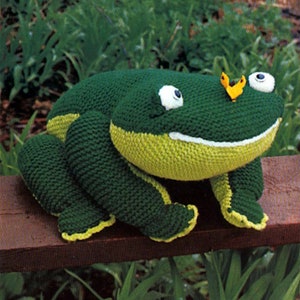 May include: A green knitted frog with yellow accents and large white eyes. The frog is sitting on a wooden surface with green grass in the background.