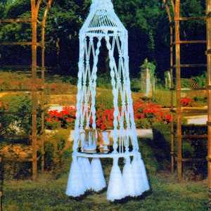 Puede incluir: Silla colgante de macramé blanco con una mesa redonda y borlas. La silla está colgada de un arco de madera en un jardín.