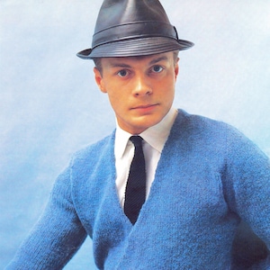 Op de afbeelding: Een man draagt een blauwe gebreide cardigan met zwarte knopen, een wit overhemd met kraag en een zwarte das. Hij draagt ook een zwarte fedora.