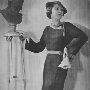 Könnte beinhalten: Eine Frau in einem schwarzen Kleid mit Gürtel und Hut, die neben einem dekorativen Ständer steht.