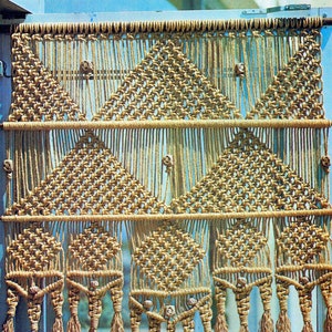 Könnte beinhalten: Ein Makramee-Wandbehang mit geometrischem Muster und Quasten. Der Wandbehang ist aus Naturfaserseil gefertigt und hat eine hellbraune Farbe.