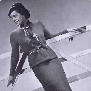 May include: A black and white photo of a woman wearing a knitted sweater and skirt set. The woman is standing in front of a wooden fence and is holding a glove in her hand.