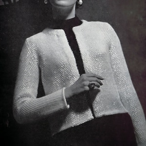 May include: A woman wearing a white knitted jacket with a black trim and a pearl earring. She is wearing a ring on her finger.