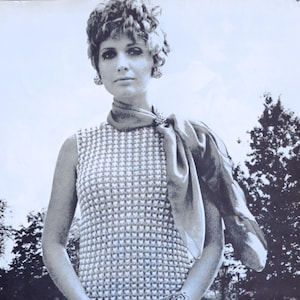 May include: A sleeveless, patterned dress with a matching scarf and bracelet. The dress has a textured, repeating pattern. The scarf is draped over the shoulders and secured with a brooch. The image is in black and white.