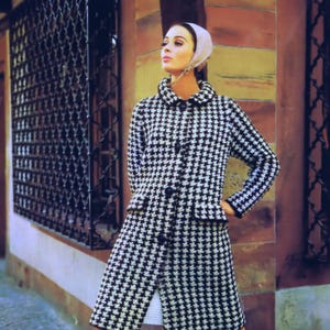 May include: A black and white houndstooth coat with a white collar and a single button fastening. The coat is worn over a white dress and black knee-high socks.