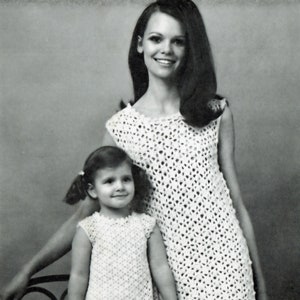 Puede incluir: Una foto en blanco y negro de una madre y su hija que llevan vestidos de crochet blanco a juego. La madre tiene el pelo largo y oscuro y está sujetando la mano de su hija. La hija tiene el pelo corto y rubio y está sonriendo.