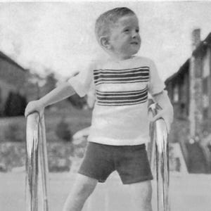 May include: A young child wearing a white t-shirt with horizontal black stripes, dark shorts, and white sneakers with black accents. The child is holding onto a metal bar, with a building in the background.