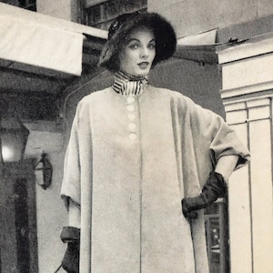 May include: A woman wearing a long, light-colored coat with a button-up front and a striped collar. She is also wearing a hat and gloves.