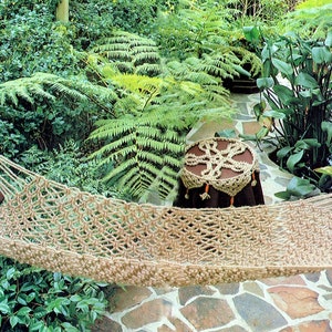 May include: A woven hammock made of natural fibers hangs between two trees in a lush green garden. The hammock is decorated with brown wooden beads and has a brown cushion.