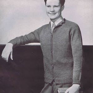 Puede incluir: Imagen en escala de grises de una persona que lleva un cárdigan gris sobre una camisa con cuello y corbata. El cárdigan tiene una parte delantera abotonada y puños acanalados. La persona sostiene libros y se apoya en una superficie oscura.