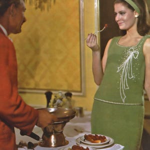 May include: A woman wearing a green dress with a white bow design is eating a small piece of food with a fork. She is seated at a table with a man in a red jacket. There is a copper fondue pot on the table with a plate of food in front of it.