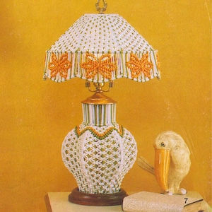 May include: A white macrame lamp base with a white and green striped shade. The shade has a border of orange butterflies. The lamp is on a table with a book and a white bird figurine.