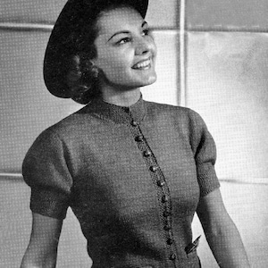 May include: A black and white photo of a woman wearing a gray knitted cardigan with a button-up front and short sleeves. She is wearing a black hat and looking up.