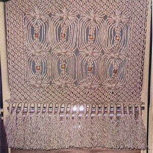 May include: A macrame wall hanging with a fringe bottom. The macrame is made with natural colored cord and features wooden beads.