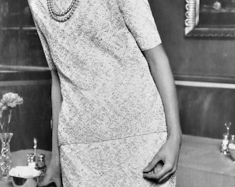 May include: A woman with short, blonde hair wearing a white, textured dress with a pearl necklace. She is standing in front of a table with a white tablecloth.