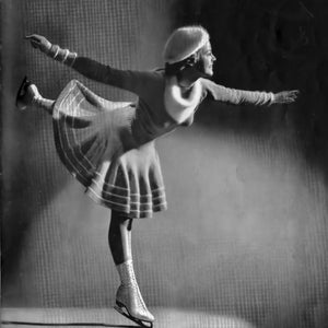 May include: A vintage black and white photograph of a figure skater in a graceful pose. She wears a light-colored sweater, skirt, and hat, with matching arm warmers and ice skates. The skater is captured mid-performance, arms outstretched, leg extended.