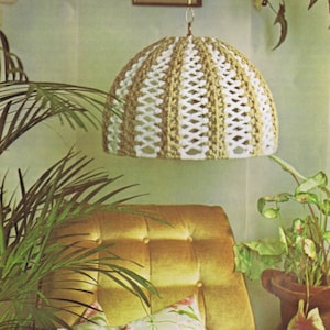 May include: A crocheted lampshade with a white and brown pattern hangs from a chain in a living room setting. The lampshade is suspended over a gold velvet sofa with two pillows. A potted plant is visible in the background.