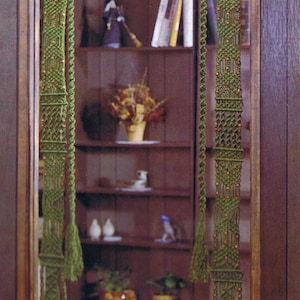Puede incluir: Una cortina de macramé verde con cuentas marrones y borlas cuelga en una puerta. El macramé está hecho con una técnica de anudado y tiene una franja en la parte inferior.