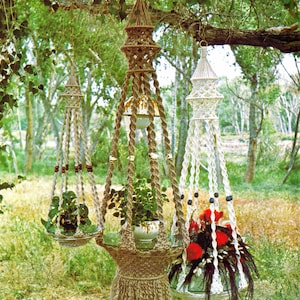 Può includere: Tre portavasi in macramè con diversi design e colori. I portavasi sono realizzati in corda naturale e presentano perline e frange. I portavasi sono appesi a un ramo d'albero e tengono piante in vaso.