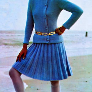 Op de afbeelding: Een vintage-stijl ensemble met een lichtblauw vest en bijpassende geplooide rok. De outfit is geaccentueerd met een gouden riem en armband. Het model draagt witte kousen en zwarte hakken. De achtergrond is een strandscène.