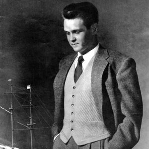 Puede incluir: Una fotografía en blanco y negro de un hombre con traje y chaleco. Está de pie con las manos en los bolsillos y mirando hacia abajo. Un barco modelo está en el fondo.