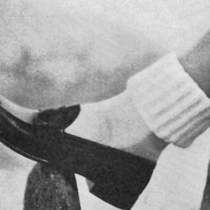 May include: Black shoe with a pointed toe, resting on a textured, dark object. A white sock with striped detailing is visible. The image is in black and white.