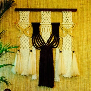 May include: A macrame wall hanging featuring a dark brown wooden rod and a woven design in cream, tan, and black. The piece has a geometric pattern with knotted details and hanging tassels, creating a bohemian aesthetic.