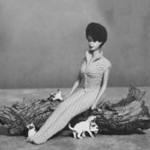 May include: A black and white photo of a doll wearing a crocheted dress sitting on a log with several small white kittens.