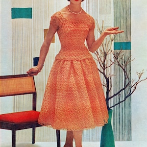 May include: A woman wearing a peach-coloured crocheted dress with a fitted bodice and a full skirt. She is standing in front of a patterned background and holding a branch.