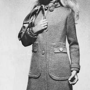 May include: A grayscale image of a woman wearing a gray, long-sleeved coat with large buttons and two front pockets. The coat has a high collar and a tailored fit. The woman is also wearing a scarf and jewelry.