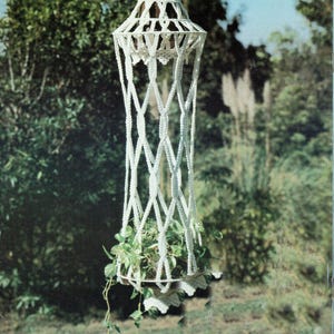 May include: A white macrame hanging planter with a geometric design, holding a green leafy plant. The planter is suspended from a metal hook, showcasing a handcrafted, bohemian style. The plant's vines cascade downwards.